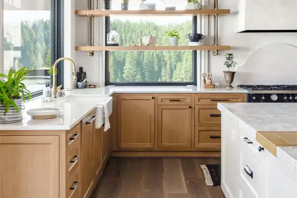 Modern kitchen with light wood cabinets, white countertops, and open shelves. A large window offers a view of trees. Features a farmhouse sink expertly installed by a plumber near me and potted plants adding a touch of nature.