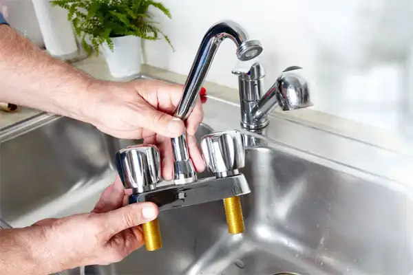 Hands holding a kitchen faucet fixture above a sink, preparing for installation. A potted plant is in the background.