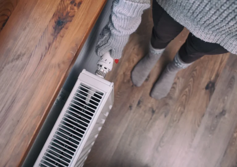 Person in a gray sweater and gloves adjusting a radiator thermostat in a room with wooden flooring.