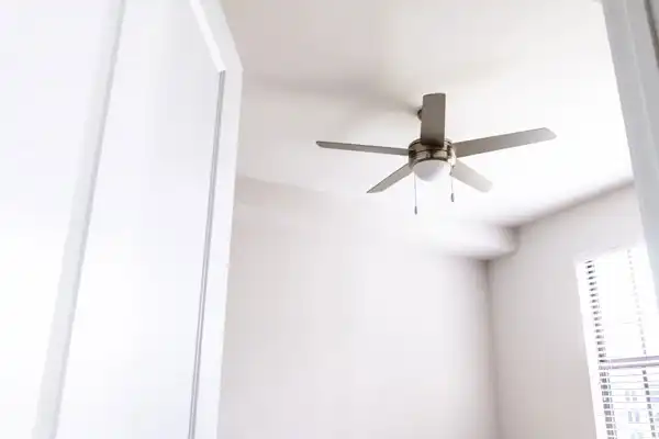 Ceiling fan with four blades in a white room with a window covered by blinds.