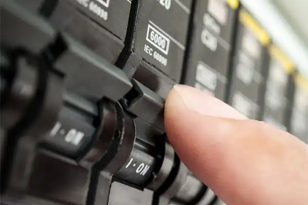 Close-up of a person’s finger turning on a switch on a black electrical circuit breaker panel marked "ON" with labels and numerical codes visible.