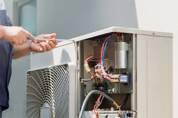 A person is repairing an outdoor air conditioning unit, with wires and components visible inside the open panel.