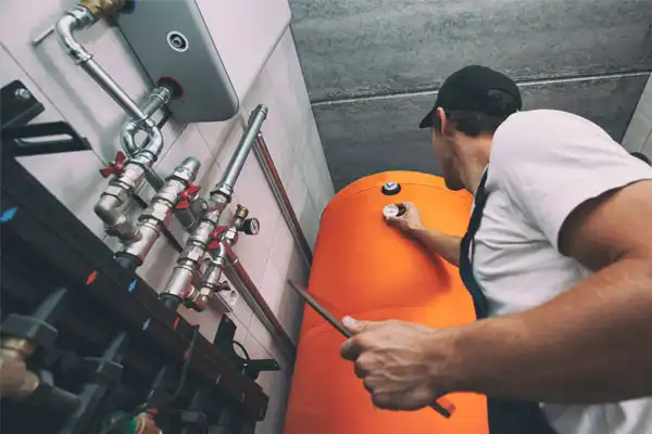 Technician in a cap and work clothes adjusting an orange boiler in a utility room with pipes and valves visible.