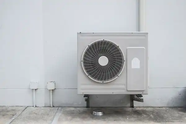 Outdoor air conditioning unit mounted on a white wall with pipes and conduit nearby.