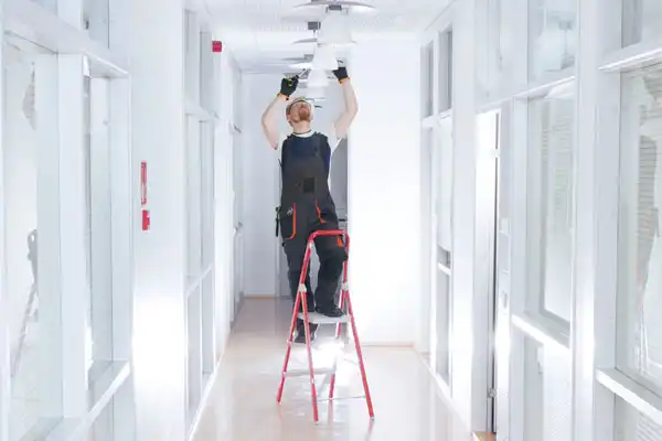 Person on a ladder installing ceiling lights in a bright, white hallway with glass partitions.