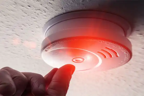 A hand is reaching toward a ceiling-mounted smoke detector with a glowing red light, suggesting it is active or being tested.