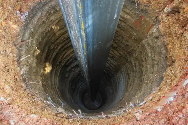 A large black pipe is being inserted vertically into a deep, circular hole dug into the ground. The surrounding earth is visible with layers of soil exposed.