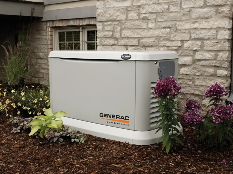 A Generac generator from the Guardian Series is installed outdoors, surrounded by flowers and mulch, against a stone wall of a house.