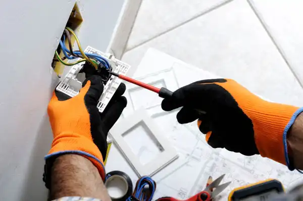 Person wearing orange gloves uses a screwdriver to fix a wall electrical outlet. Nearby, tools and wiring components are scattered on the floor.