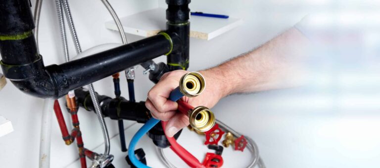 A person is holding two hoses with brass fittings in front of various plumbing pipes and valves.