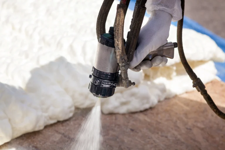 A person wearing a glove is using a spray gun to apply white foam insulation on a surface.